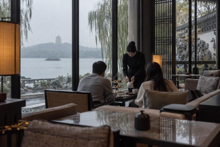 Two guests share a tiered afternoon tea stand by tall rain-streaked windows in a West Lake hotel lounge in Hangzhou while a server pours tea, with willows and a grey lake beyond.