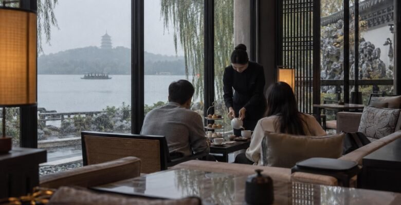 Two guests share a tiered afternoon tea stand by tall rain-streaked windows in a West Lake hotel lounge in Hangzhou while a server pours tea, with willows and a grey lake beyond.
